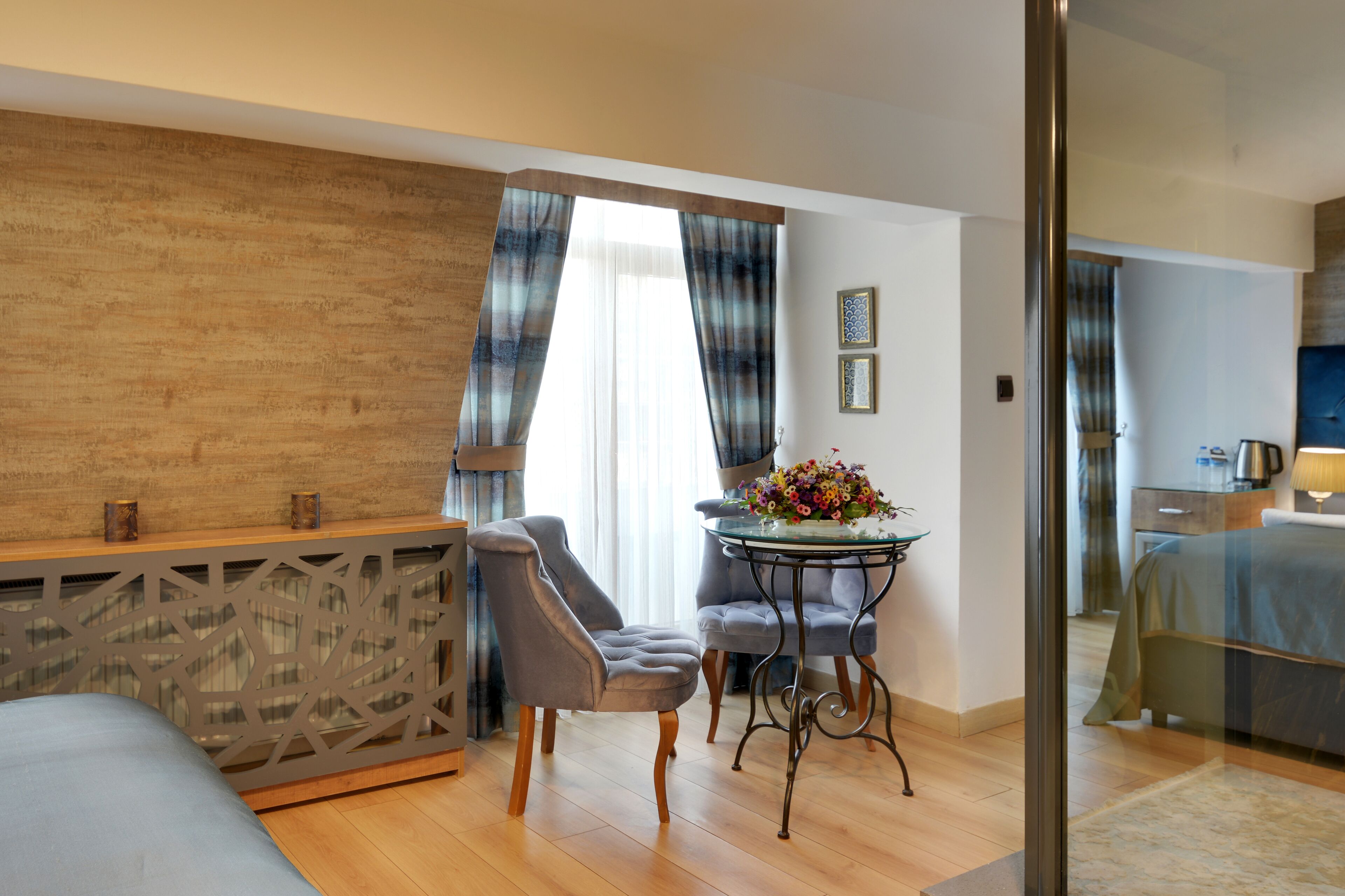 A modern hotel room with a wooden accent wall, a dining area with a table and chairs, and a separate bedroom with a bed and nightstand.