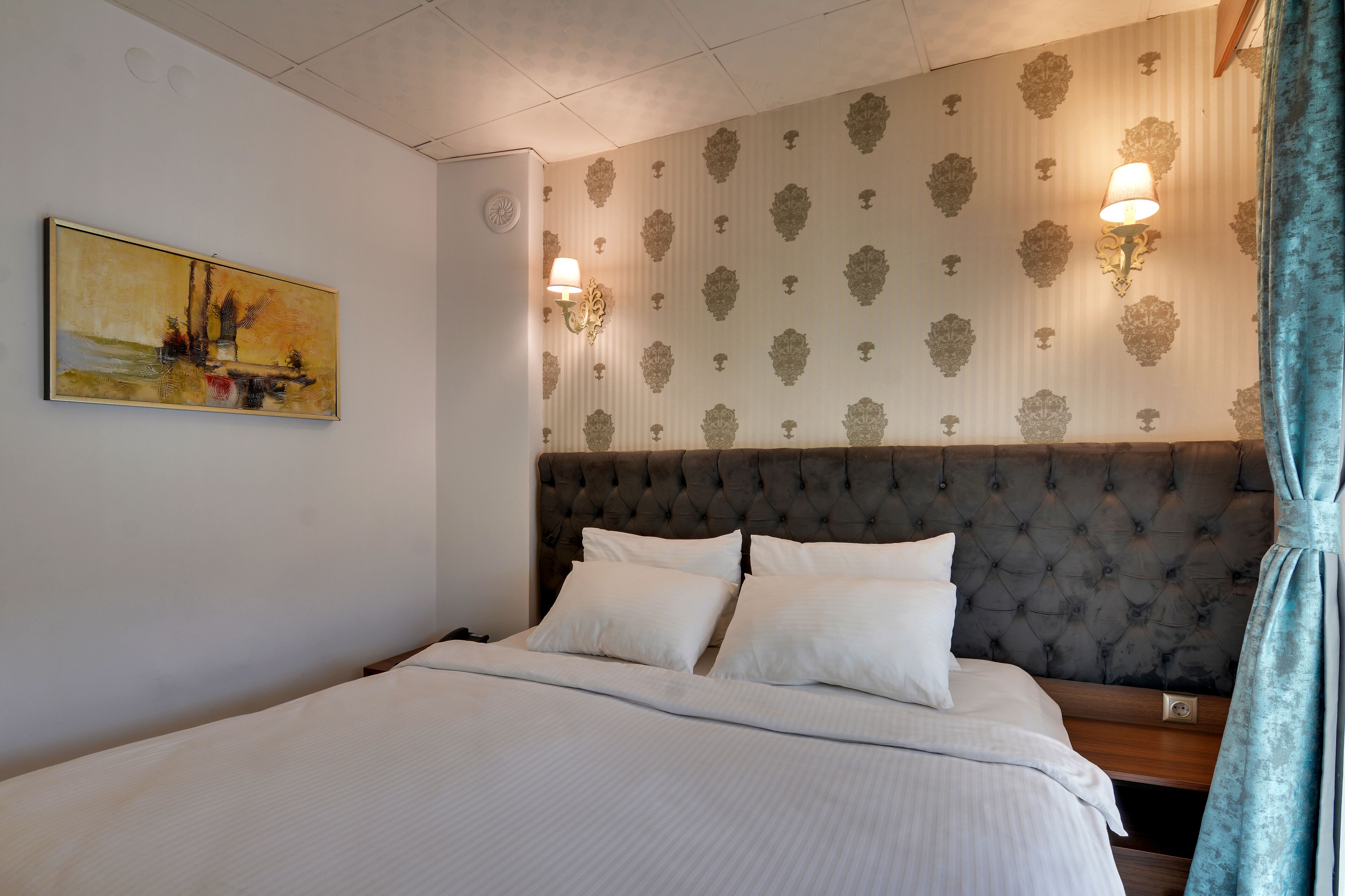 A neatly made bed with a tufted headboard, two wall-mounted lamps, and a patterned wallpaper.