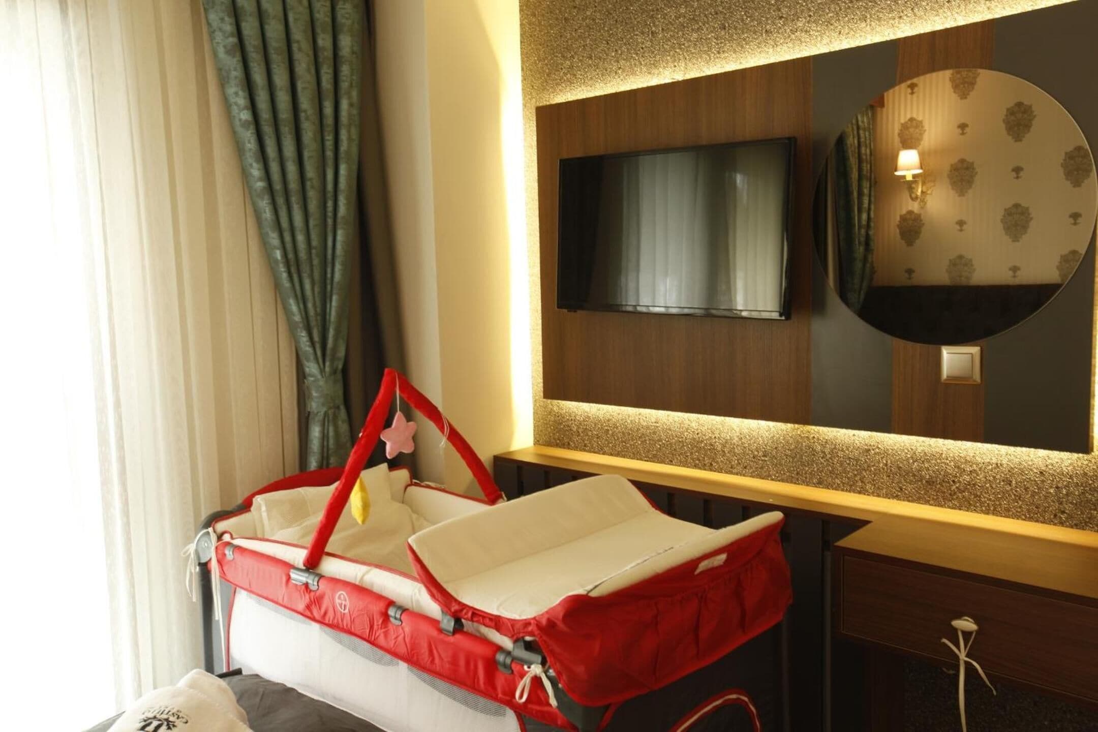 Ein Hotelzimmer mit einem roten Kinderwagen, einem flachbildfernseher an der Wand und einem runden Spiegel mit einem gemusterten Rahmen.