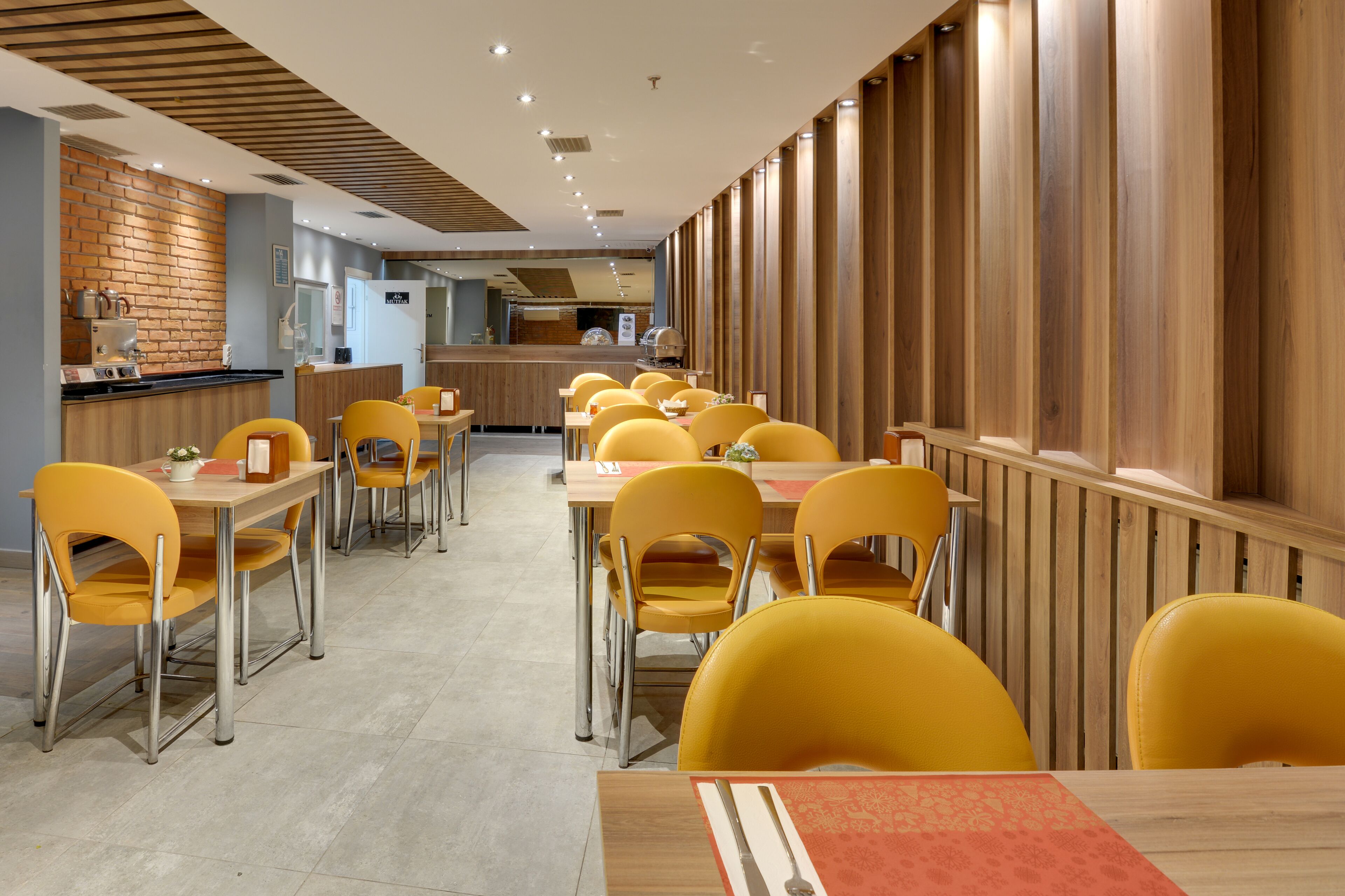 A restaurant with yellow chairs and tables, wooden paneling on the walls, and a kitchen area in the background.