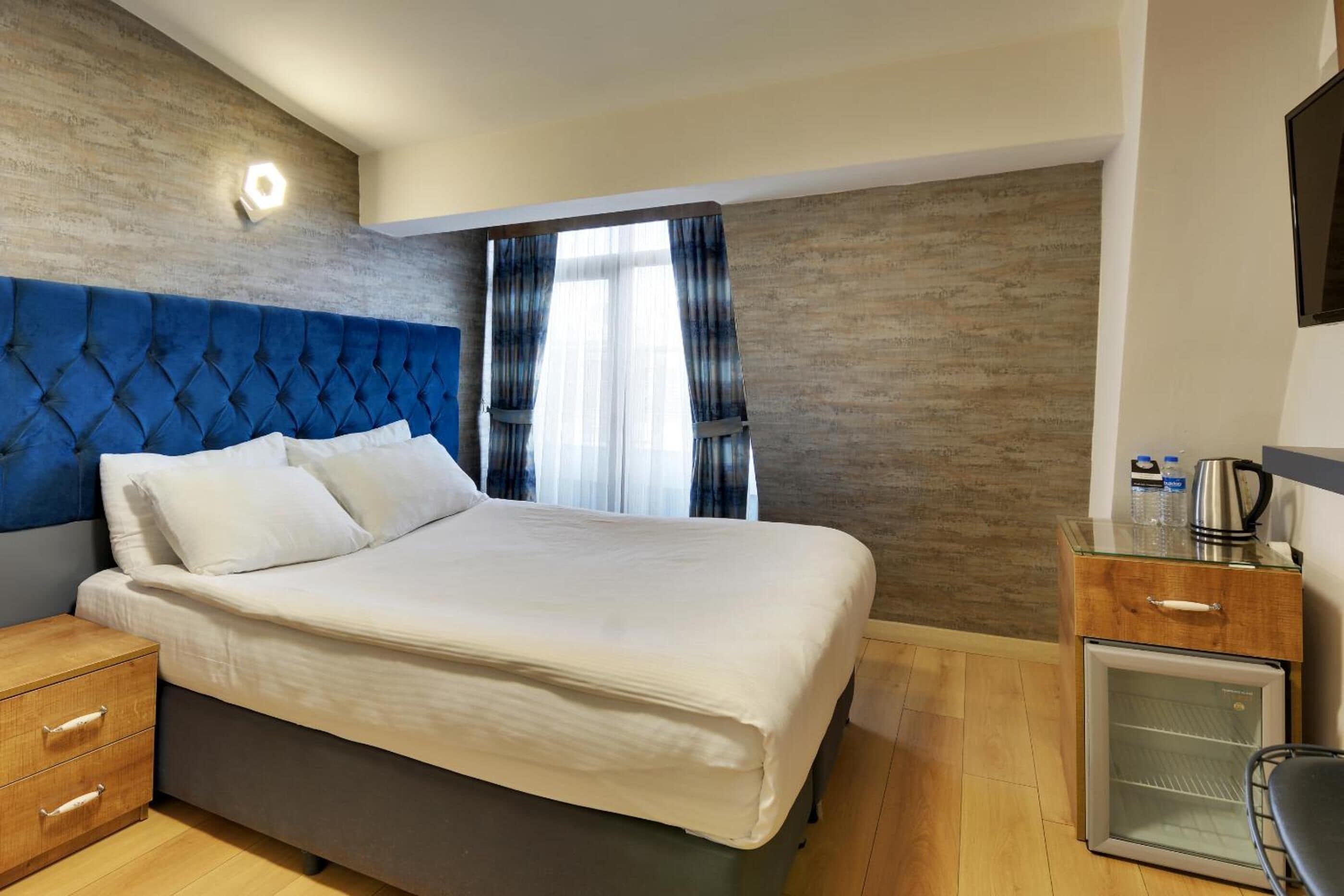 A hotel room with a large bed, a wooden nightstand, a small refrigerator, and a window with curtains.