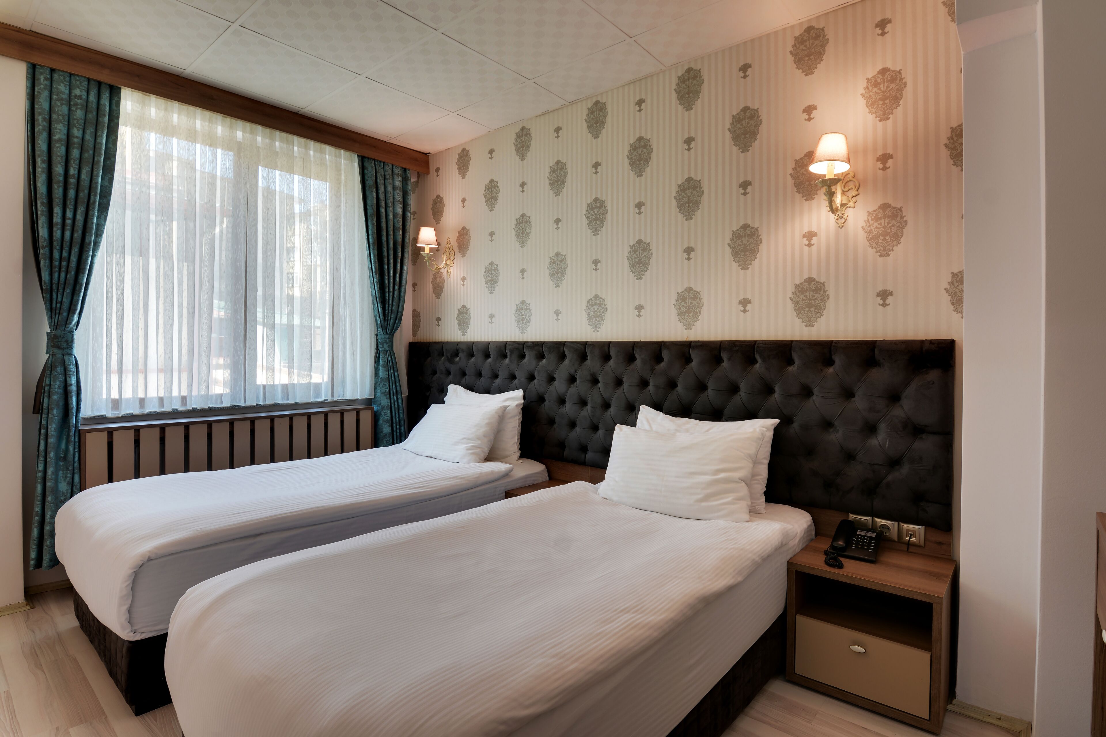 Habitación de hotel con dos camas individuales, cabecero tapizado oscuro, mesita de noche con lámpara y papel tapiz estampado.