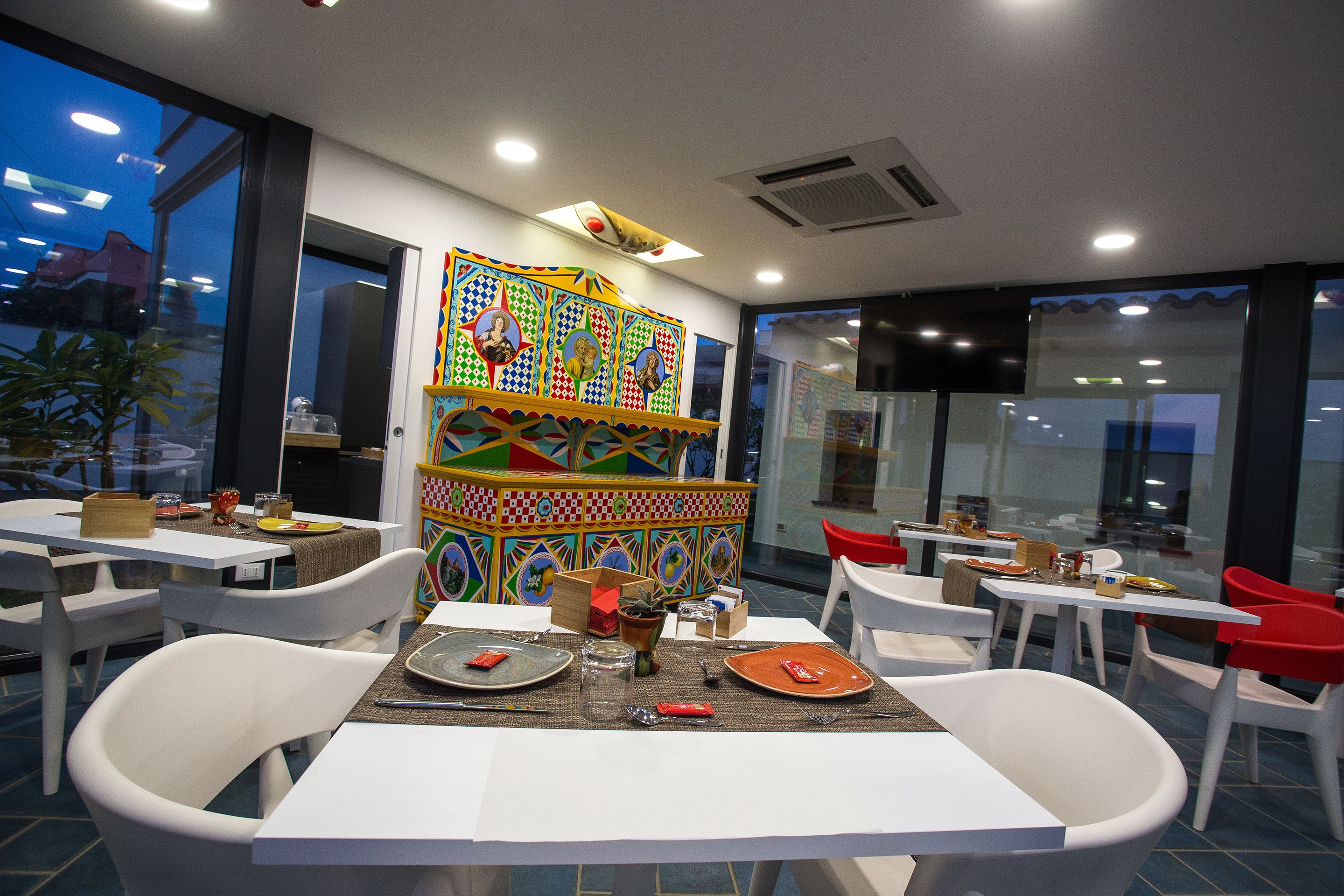 Un restaurant avec des tables et des chaises blanches, une fresque murale colorée et une vue sur la ville au crépuscule.