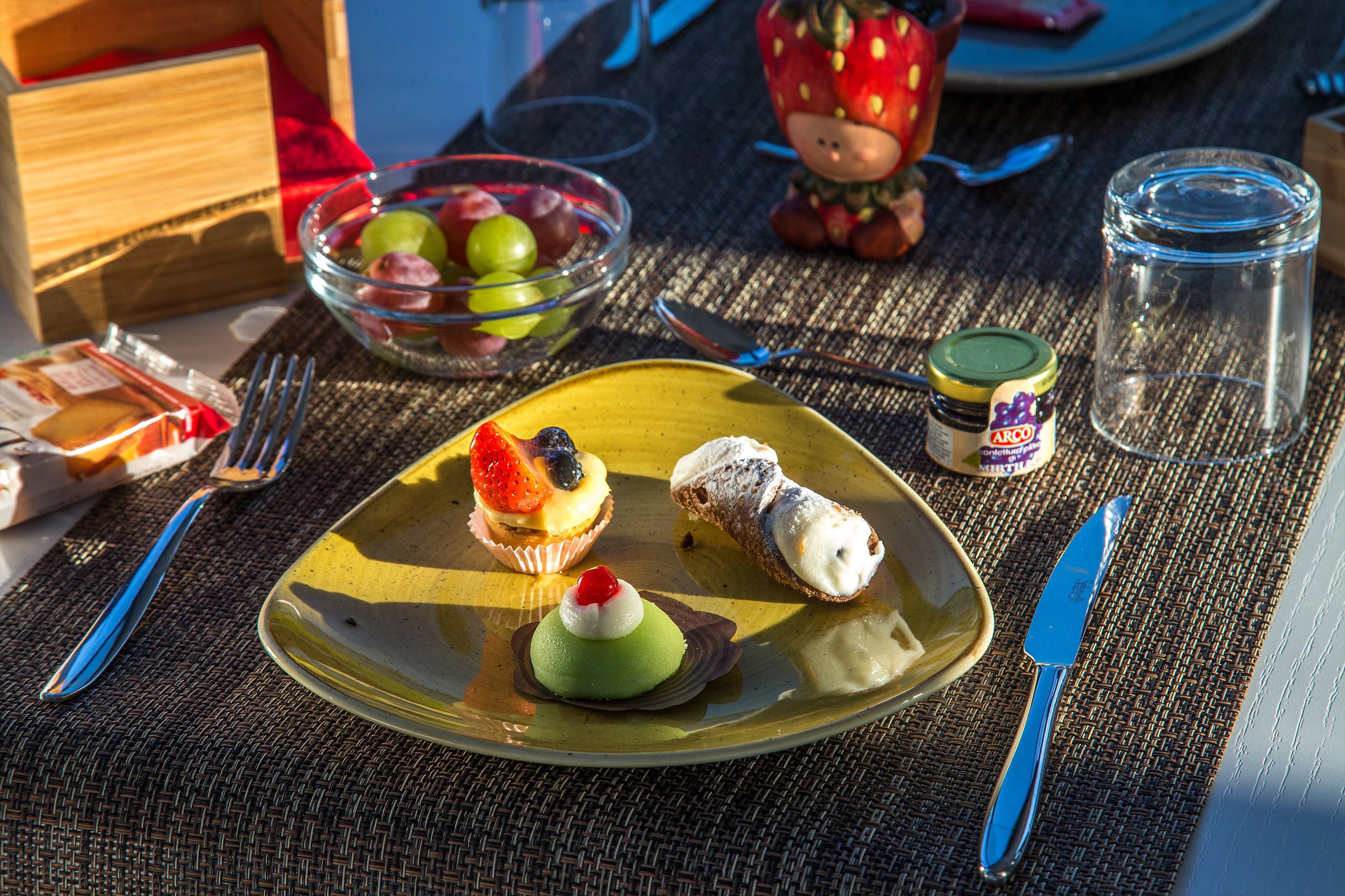 Uma mesa posta com uma travessa de sobremesas, uma cesta de frutas e uma pequena estatueta.