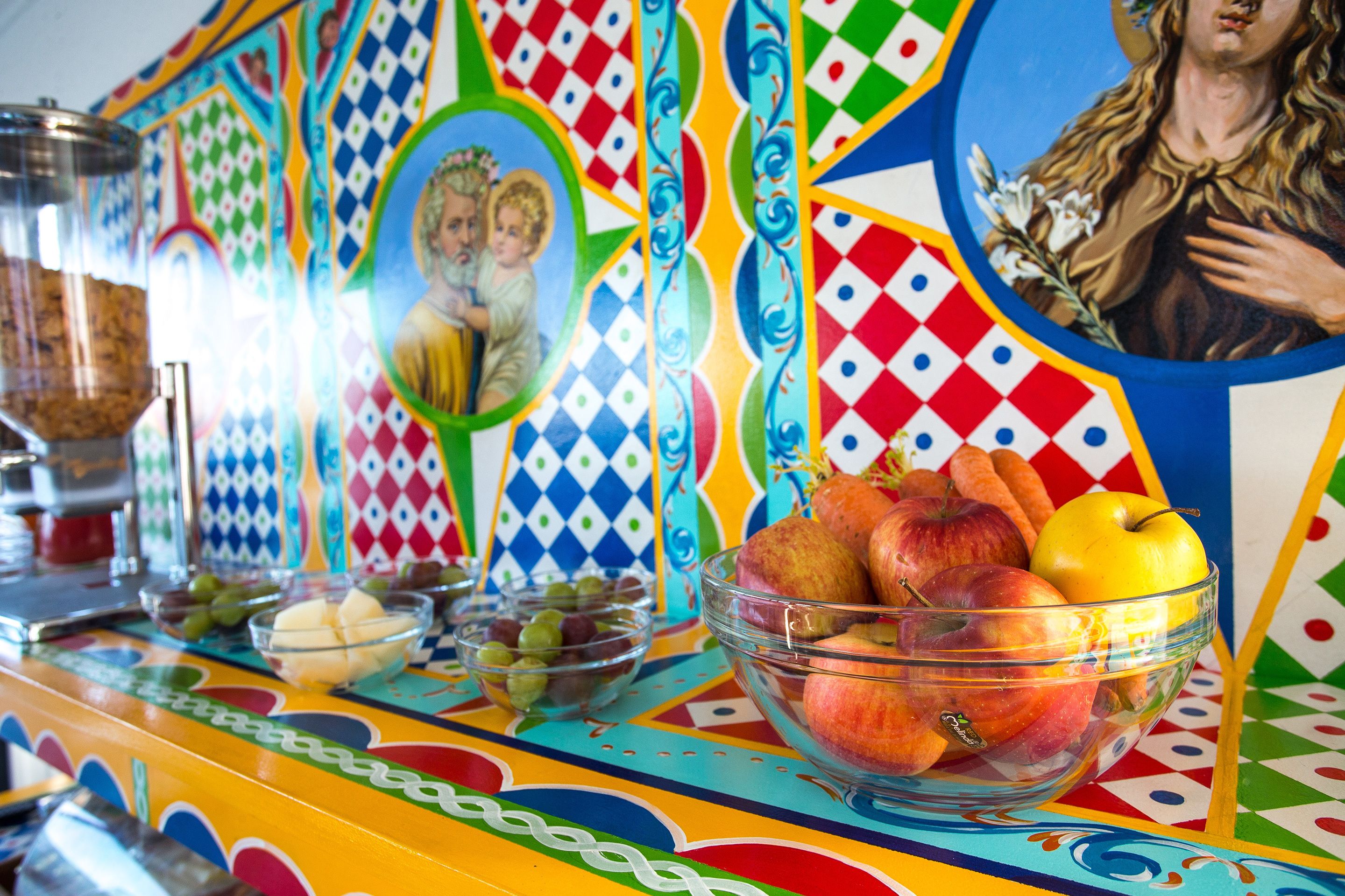 Un mural colorido con una figura religiosa, un cuenco de frutas y un recipiente de vidrio sobre una mesa.