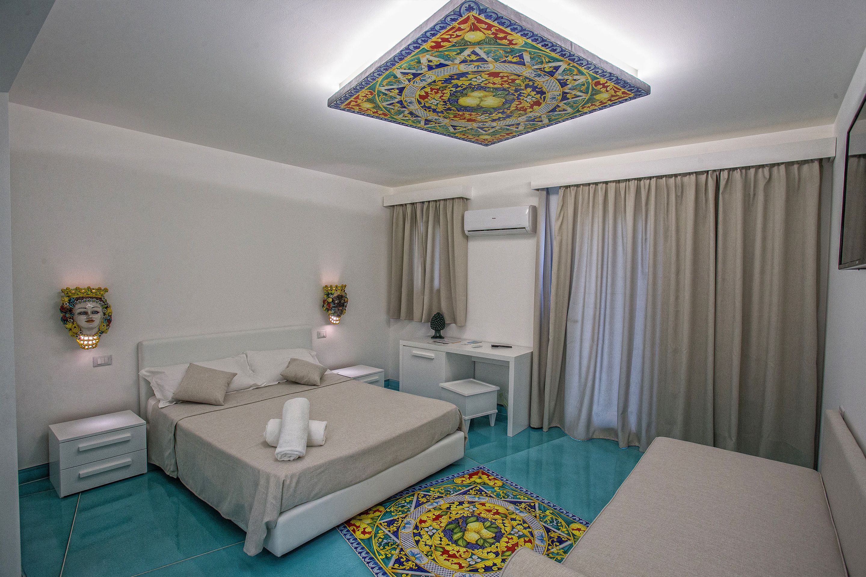 Un dormitorio con cama, mesitas de noche, escritorio, unidad de aire acondicionado y techo con diseño decorativo.