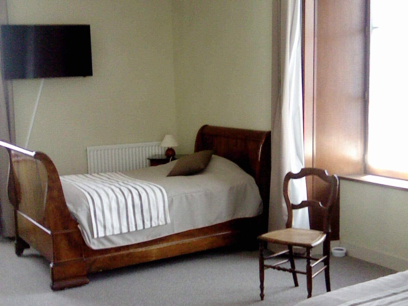 Un dormitorio con cama de madera, una silla, una mesita de noche con lámpara y un televisor en la pared.
