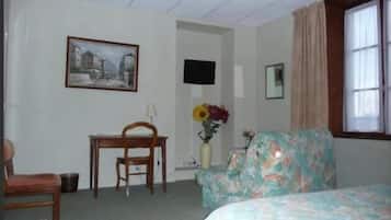 A hotel room with a bed, a chair, a desk, a television, a painting, a vase with flowers, and a window with curtains.