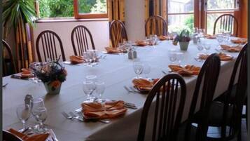 A long dining table set for a meal with chairs around it, large windows with a view of greenery, and a potted plant on the table.
