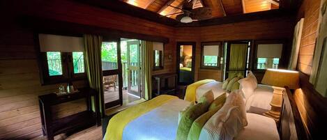 A wooden cabin bedroom with a large bed, a desk, and a balcony with a view of the greenery outside.