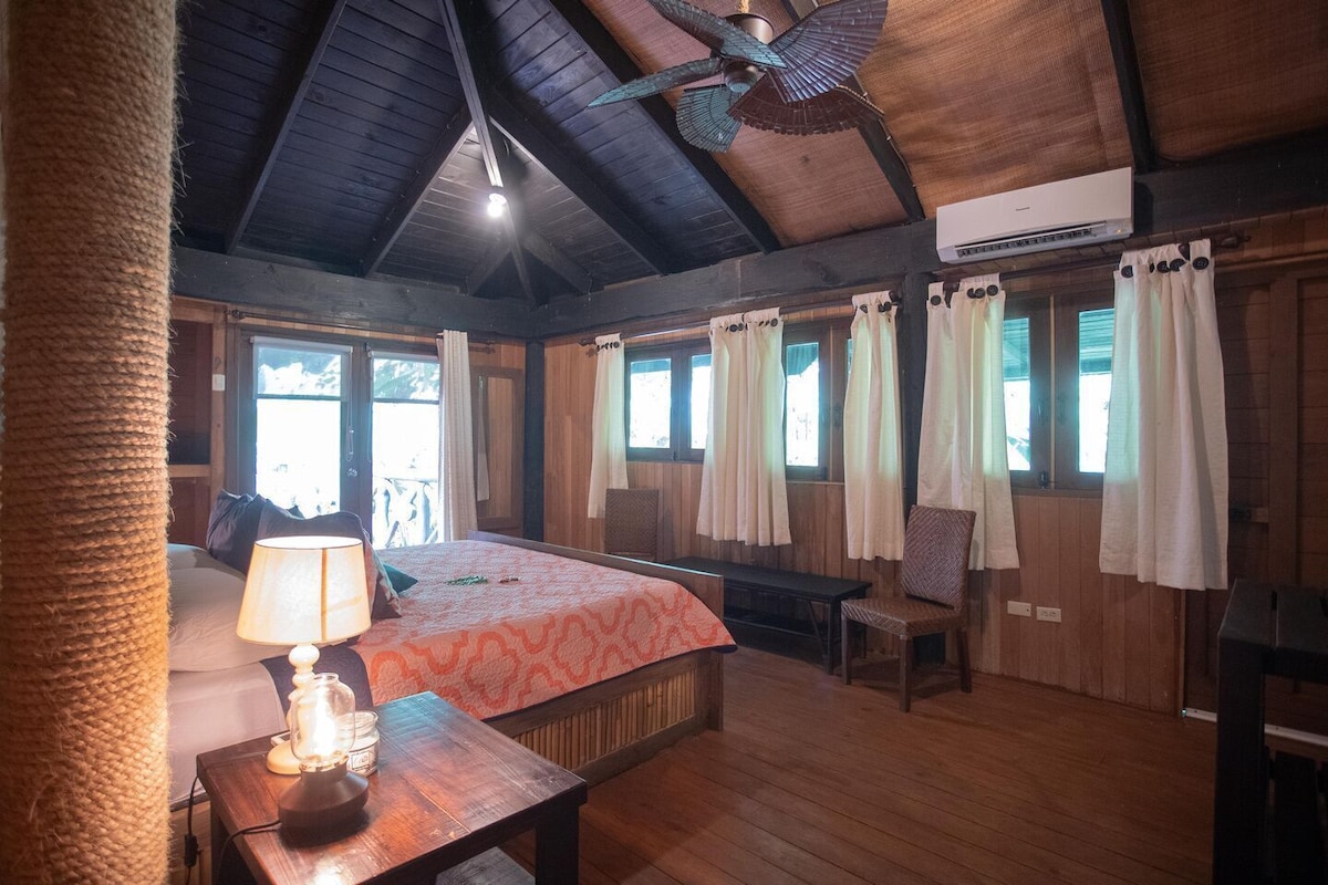Una habitación de madera con una cama, una mesa con una lámpara, una silla y un ventilador de techo.