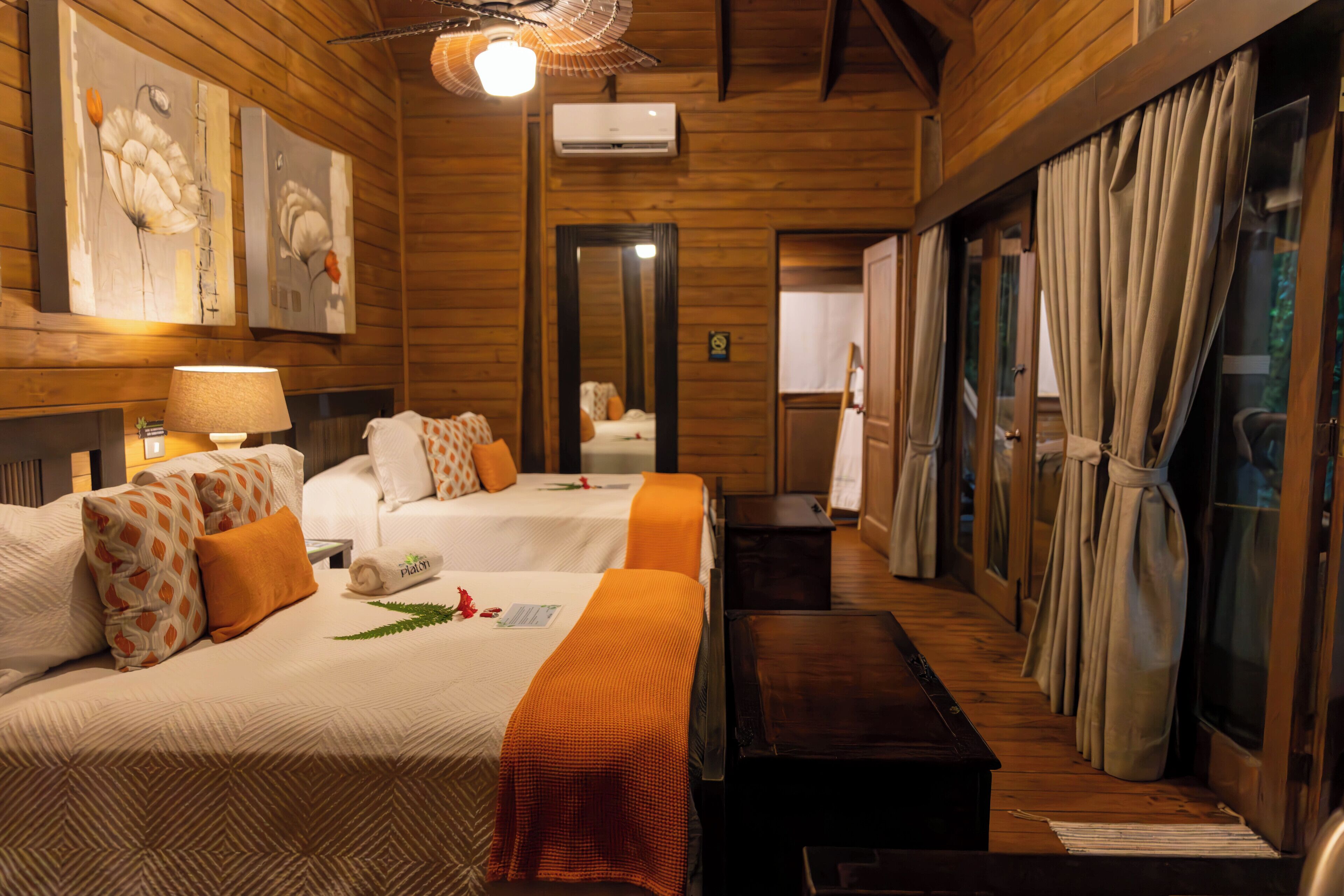 Habitación de hotel con dos camas, piso de madera, ventilador de techo y obras de arte en las paredes.