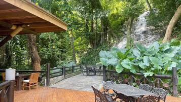 Una terraza de madera al aire libre con una mesa y sillas, rodeada de exuberante follaje y un sendero de piedra.