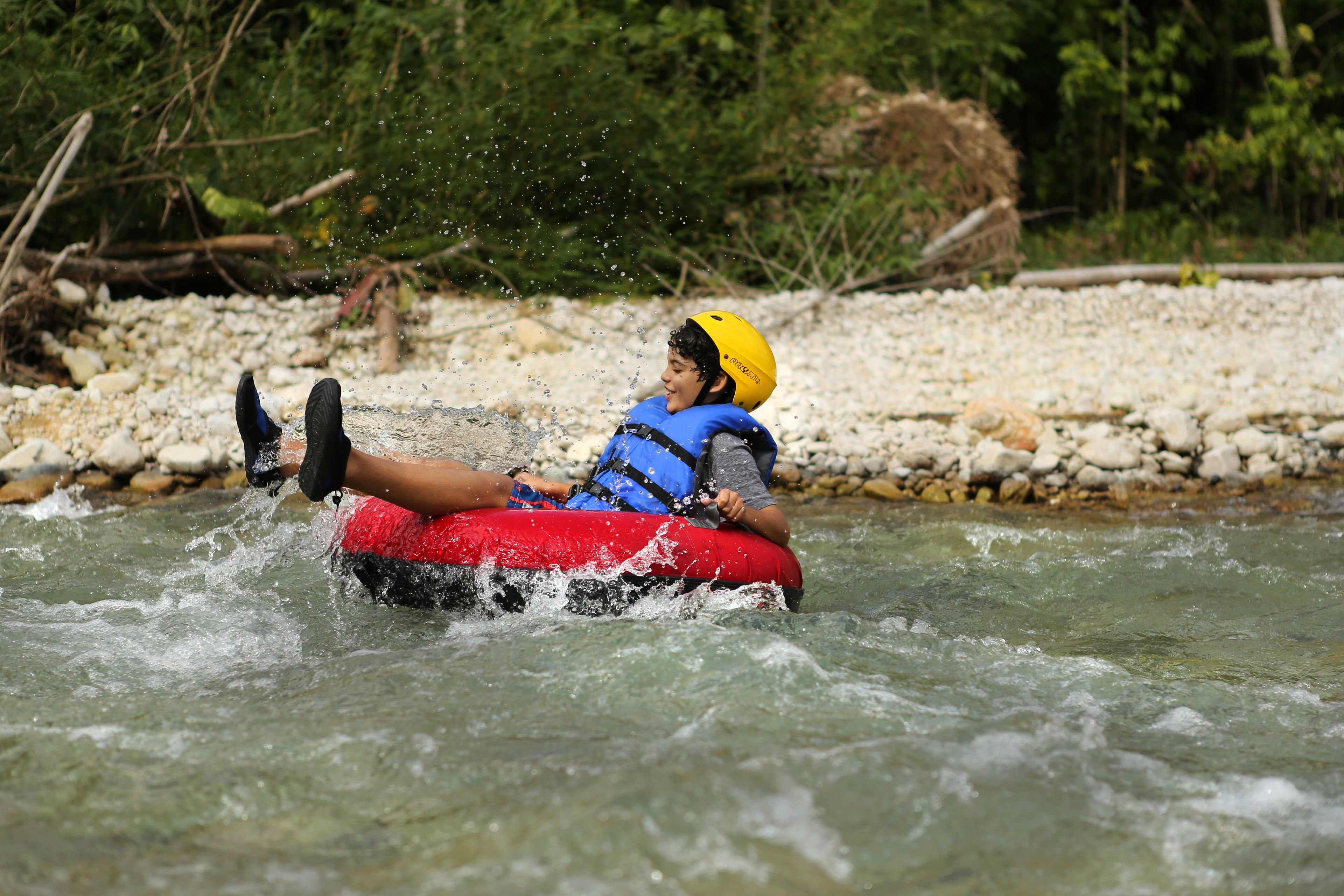 A person in a red raft with a yellow helmet, wearing a blue life jacket, floating on a river surrounded by rocks and greenery.