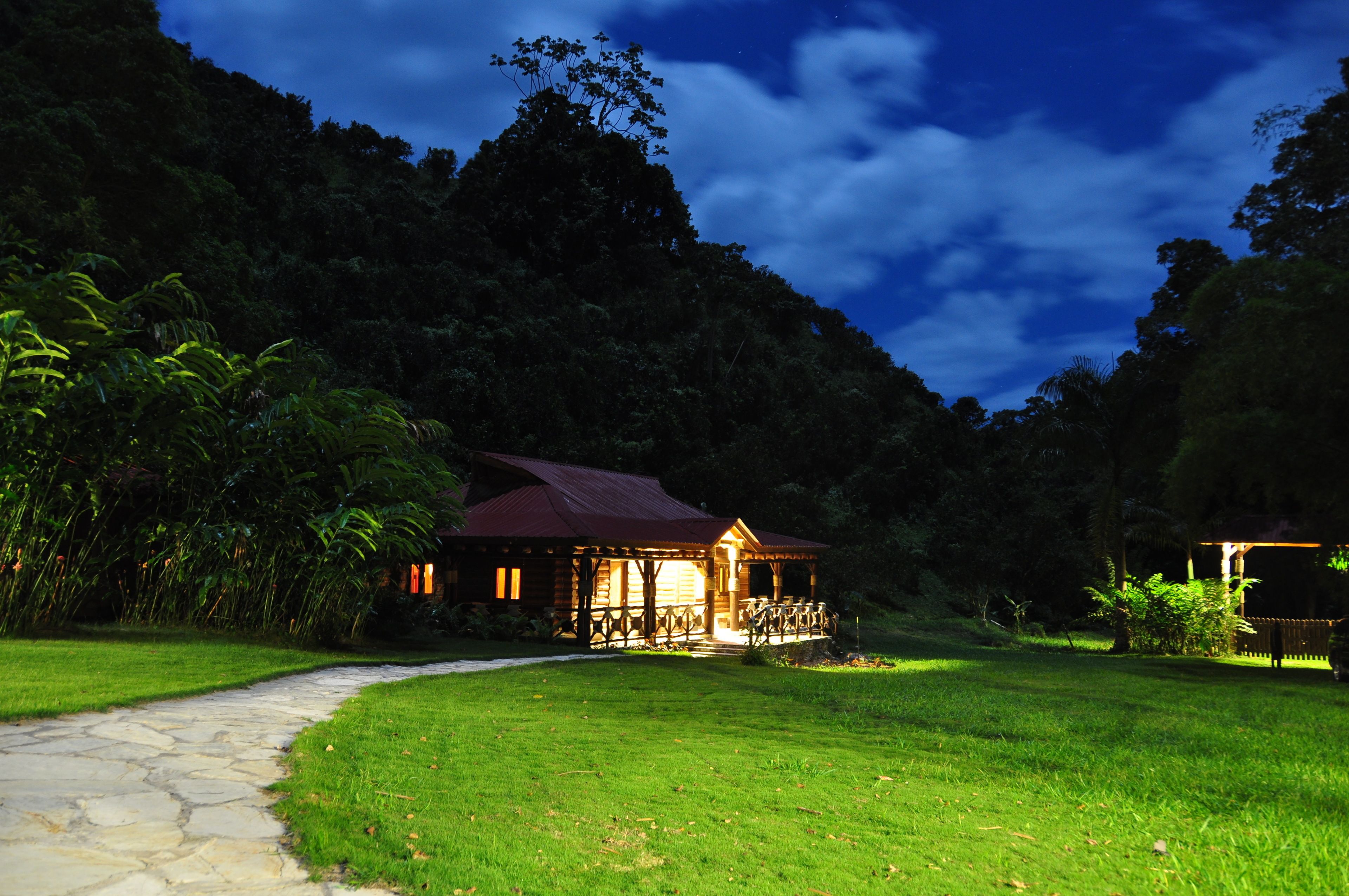 A wooden cabin with a porch, surrounded by lush greenery and a stone pathway leading to it.