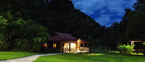 A wooden cabin with a porch, surrounded by lush greenery and a stone pathway leading to it.