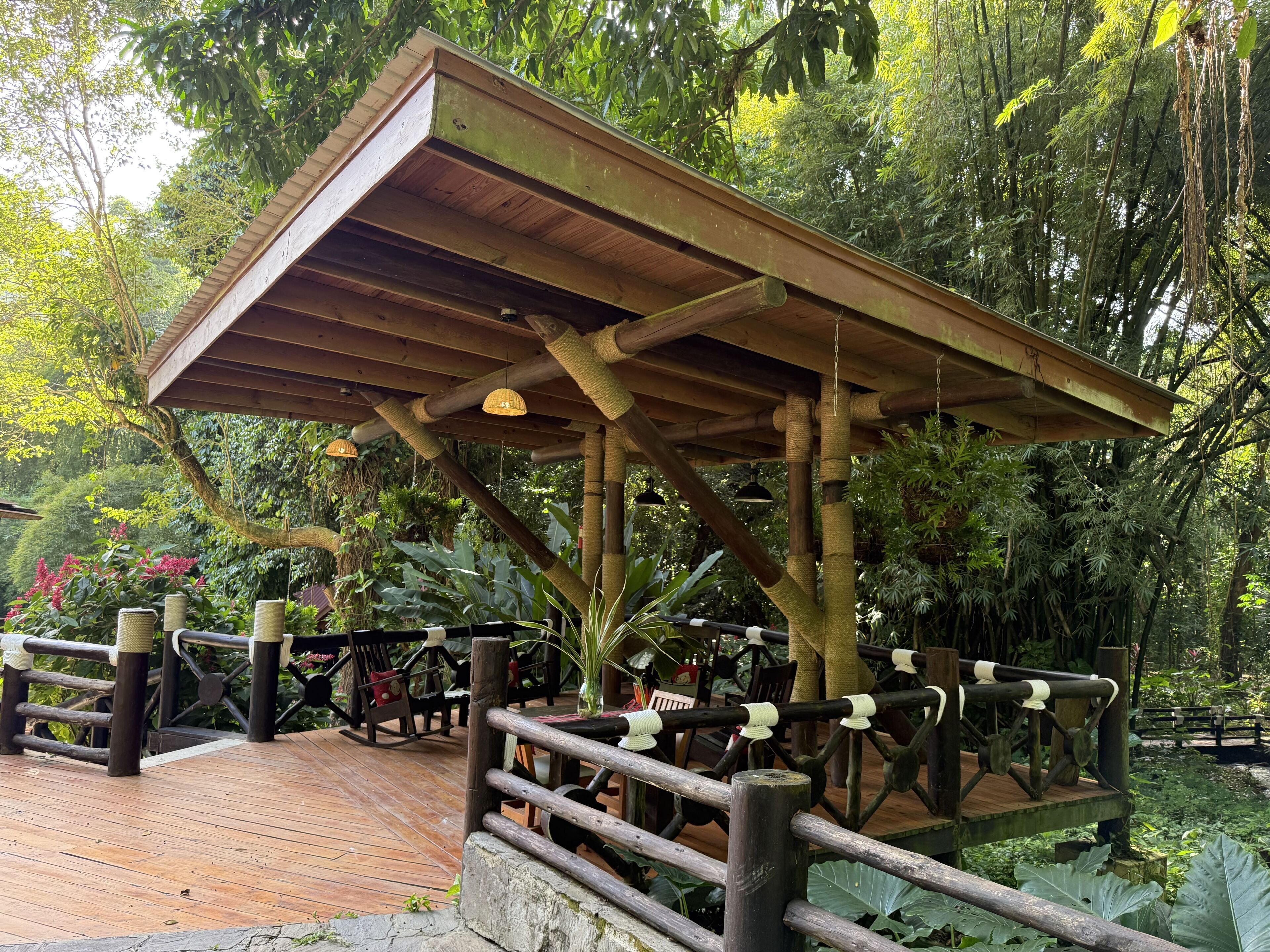 A wooden pavilion with a sloped roof, surrounded by lush greenery and a wooden deck with seating.