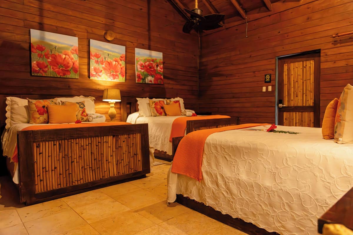 Una habitación de madera con dos camas, cada una con ropa blanca de cama y almohadones naranjas. Hay cuadros con motivos florales en la pared, un ventilador de techo y una puerta con panel de bambú.