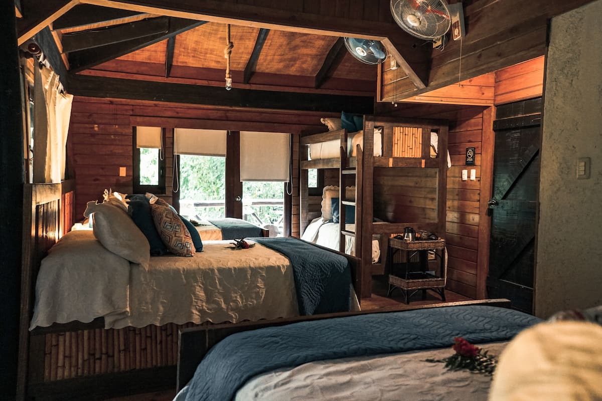 Interior rústico de una cabaña de madera con una cama, una mesita y un ventilador.