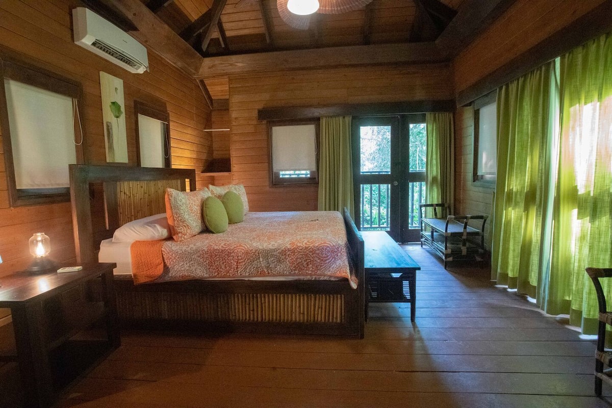 Un dormitorio de estilo cabaña con cama, mesitas de noche, un banco y una ventana con cortinas.