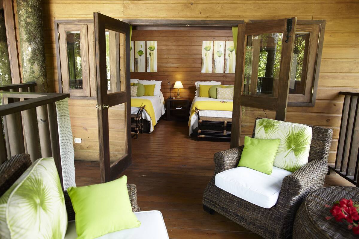 Una cabaña de madera con dos camas, una mesita de noche, muebles de mimbre y vistas al exterior.