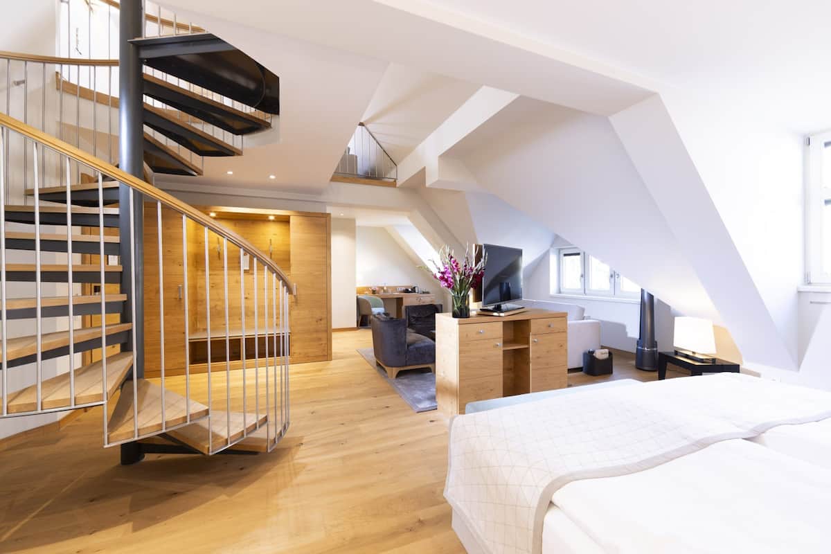 A modern loft bedroom with a wooden staircase, a bed, a desk, and a TV.
