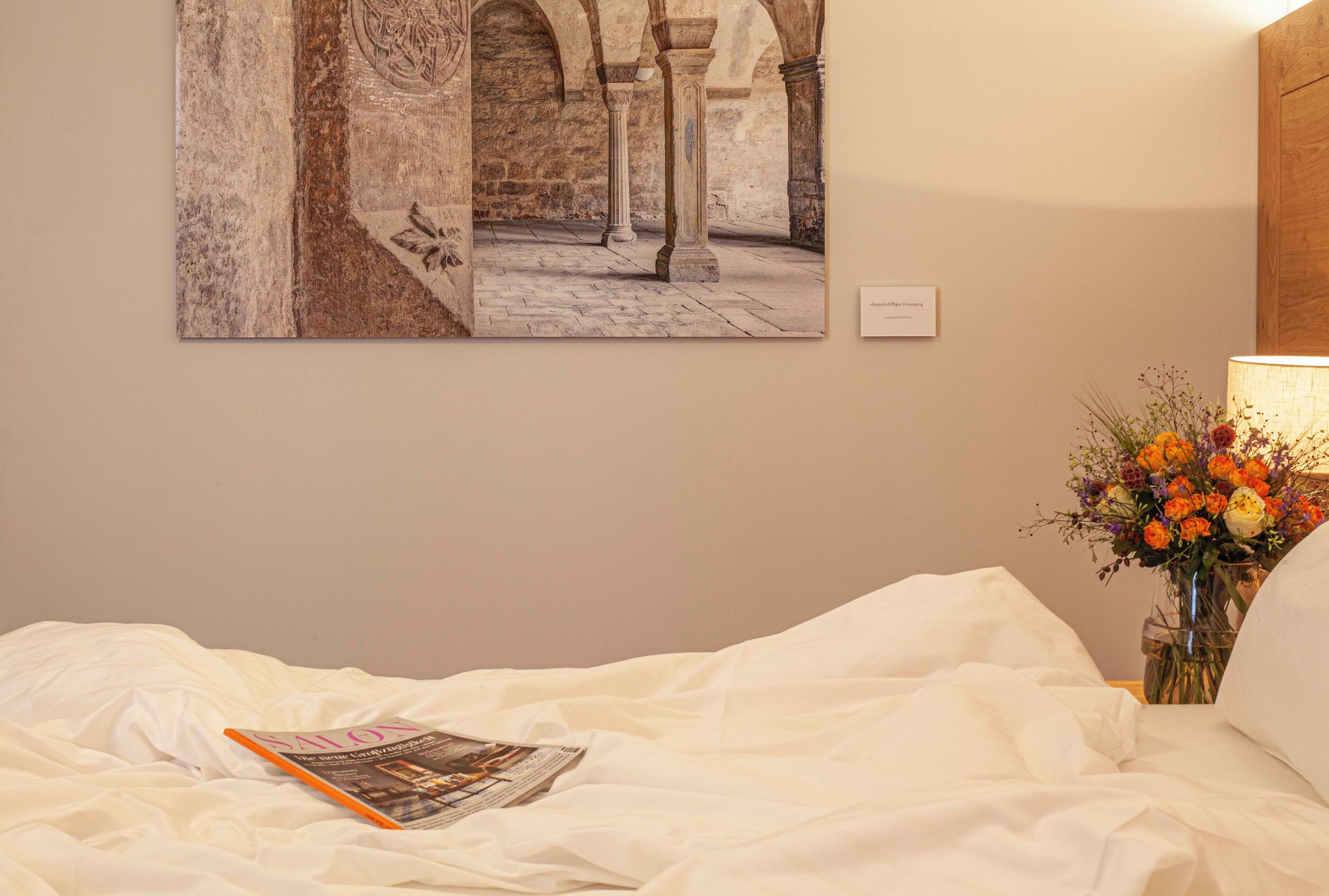 A hotel room with a bed, a framed picture on the wall, a magazine on the bed, and a vase with flowers on the nightstand.