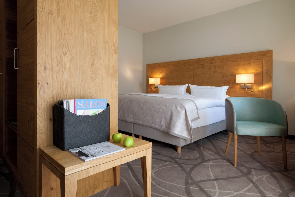 A hotel room with a wooden headboard, two bedside lamps, a bed with white linens, a small table with a magazine and apples, and a teal chair.