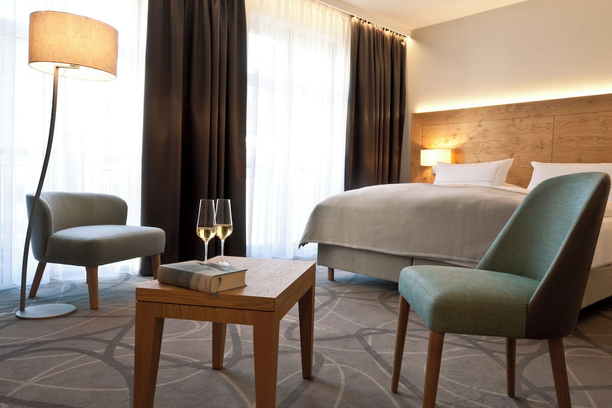 A hotel room with a bed, two armchairs, a table with a book and glasses, and a lamp.