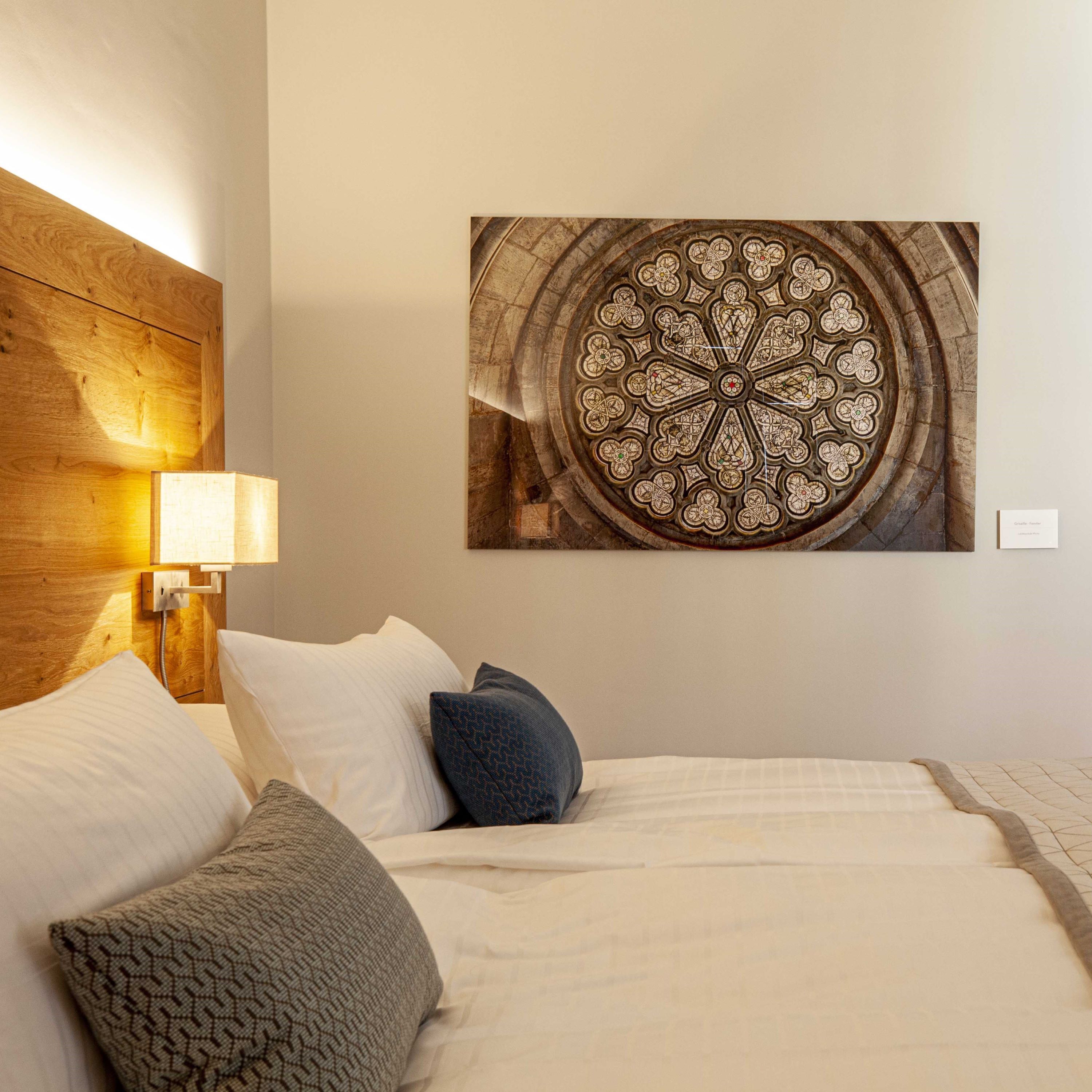 A hotel room with a bed, pillows, a wooden headboard, a wall-mounted lamp, and a large circular stained glass window.
