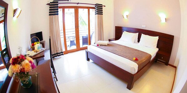 A hotel room with a large bed, a desk with a computer and a vase of flowers, a sliding glass door to the balcony, and wall-mounted lights. - Hotel Shanelo (Negombo)