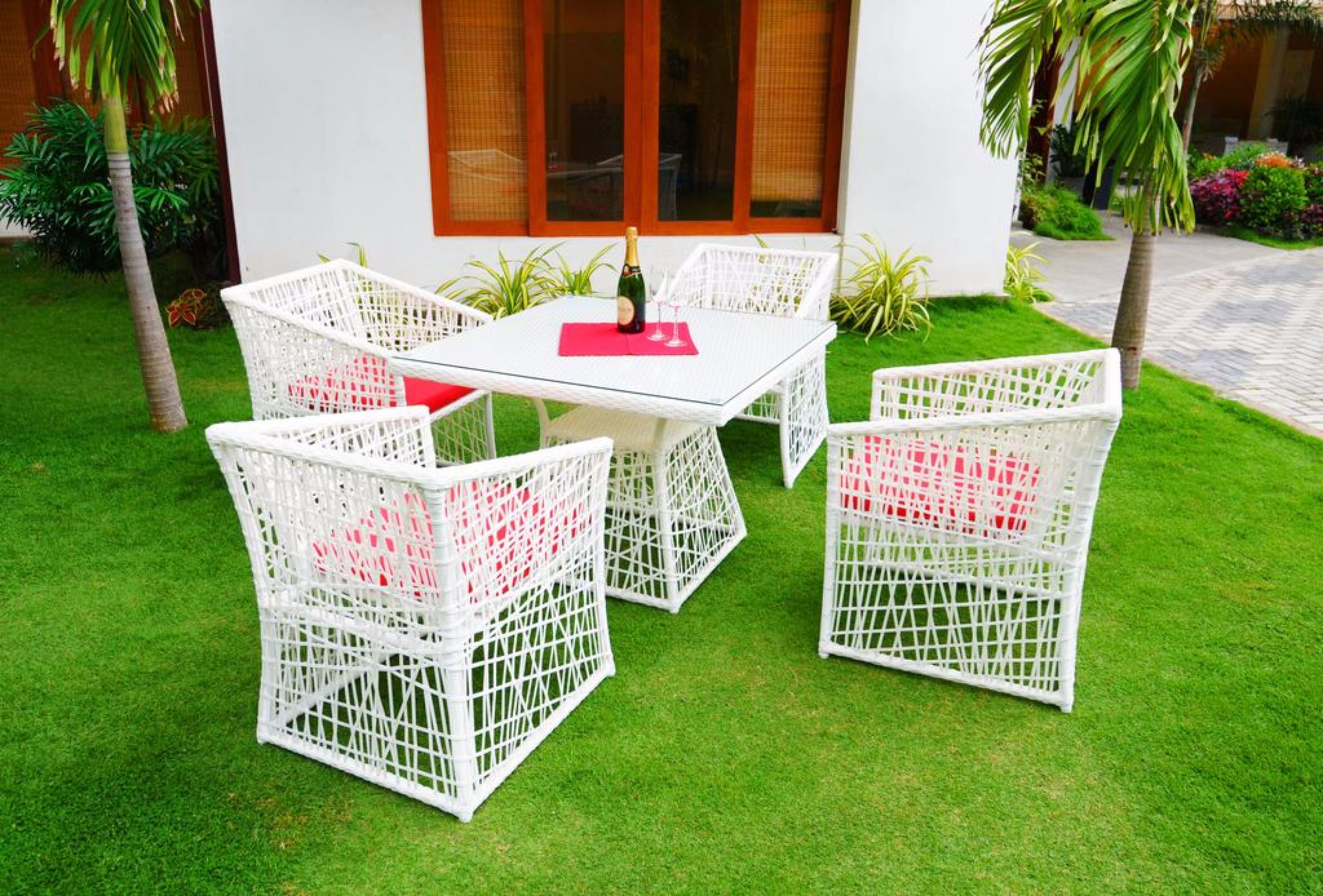 A white wicker outdoor seating area with a glass table, a bottle of champagne, and red cushions.