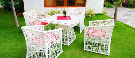 A white wicker outdoor seating area with a glass table, a bottle of champagne, and red cushions.