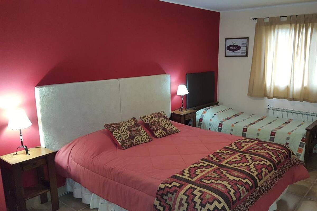 A bedroom with a large bed, two bedside tables, a desk, a chair, a window with curtains, and a wall-mounted TV.