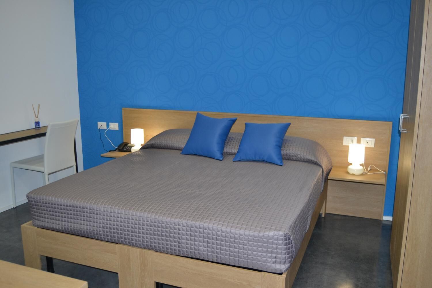 Cama bem arrumada com colcha cinza, duas almofadas azuis e cabeceira de madeira. Há abajures de cada lado da cama.