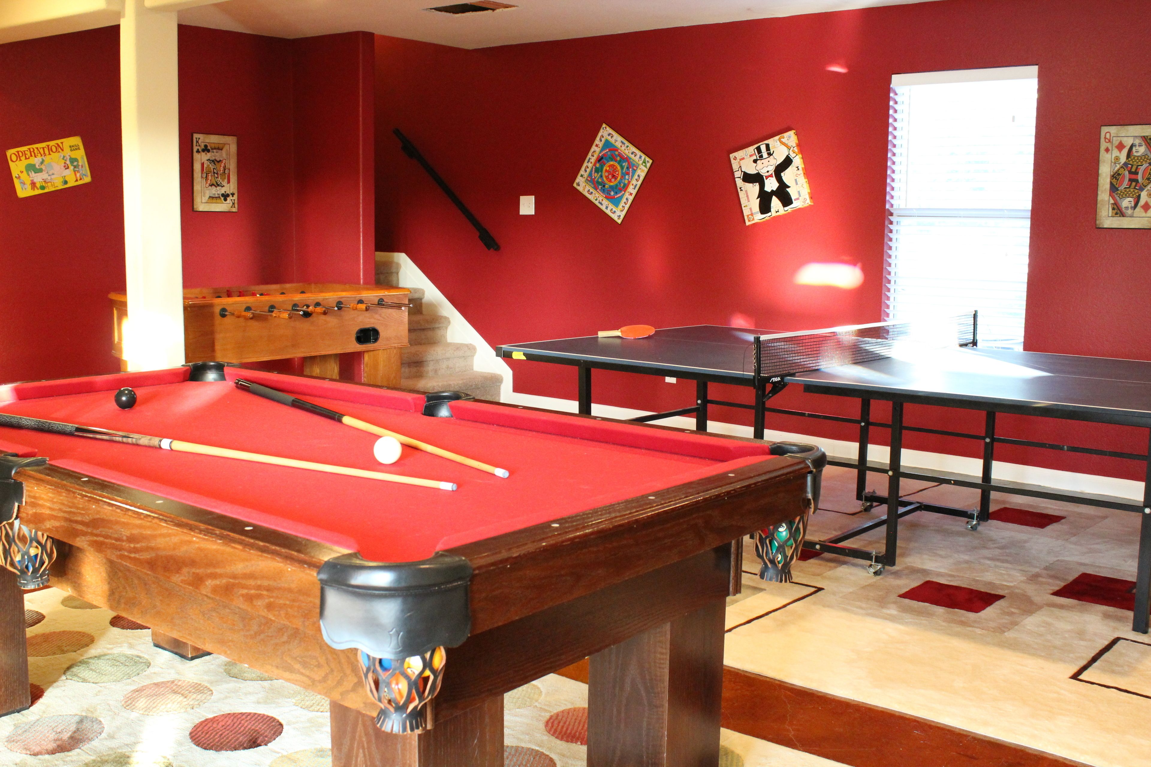 Una mesa de billar con superficie de fieltro rojo, una mesa de fútbol de mesa y una mesa de ping pong en una habitación con paredes rojas y una escalera.