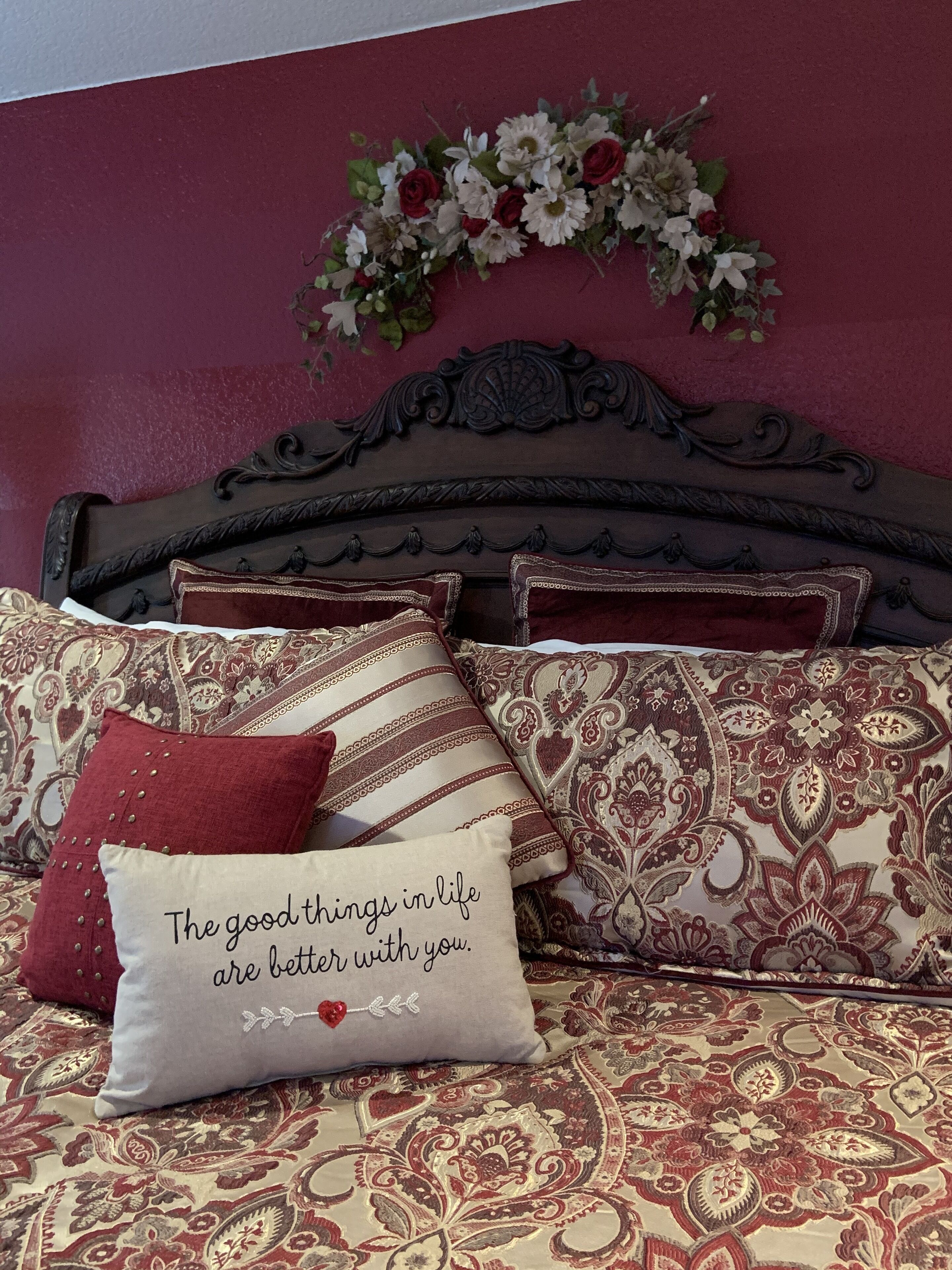 Un dormitorio con colcha estampada, varias almohadas, un arreglo floral decorativo sobre la cabecera y una pared pintada de color rojo intenso.