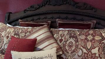 Un dormitorio con colcha estampada, varias almohadas, un arreglo floral decorativo sobre la cabecera y una pared pintada de color rojo intenso.
