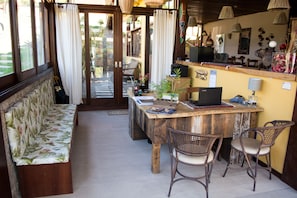 A cozy room with a wooden bench, wicker chairs, a wooden table with a laptop, and a small garden visible through the door. - Pousada Gaia (Imbituba)