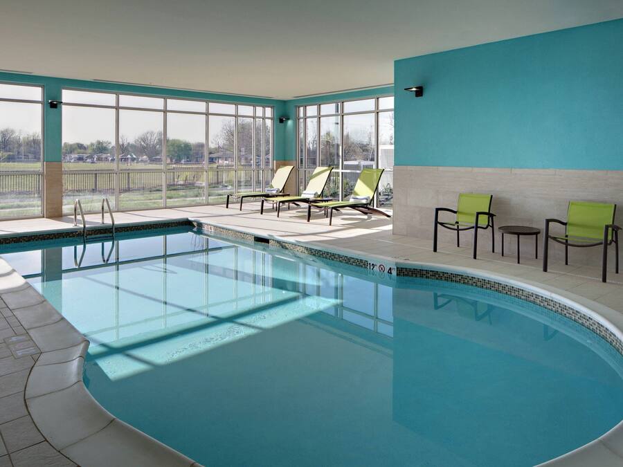 An indoor swimming pool with clear blue water, sunlit by large windows, and furnished with lounge chairs and a small table.