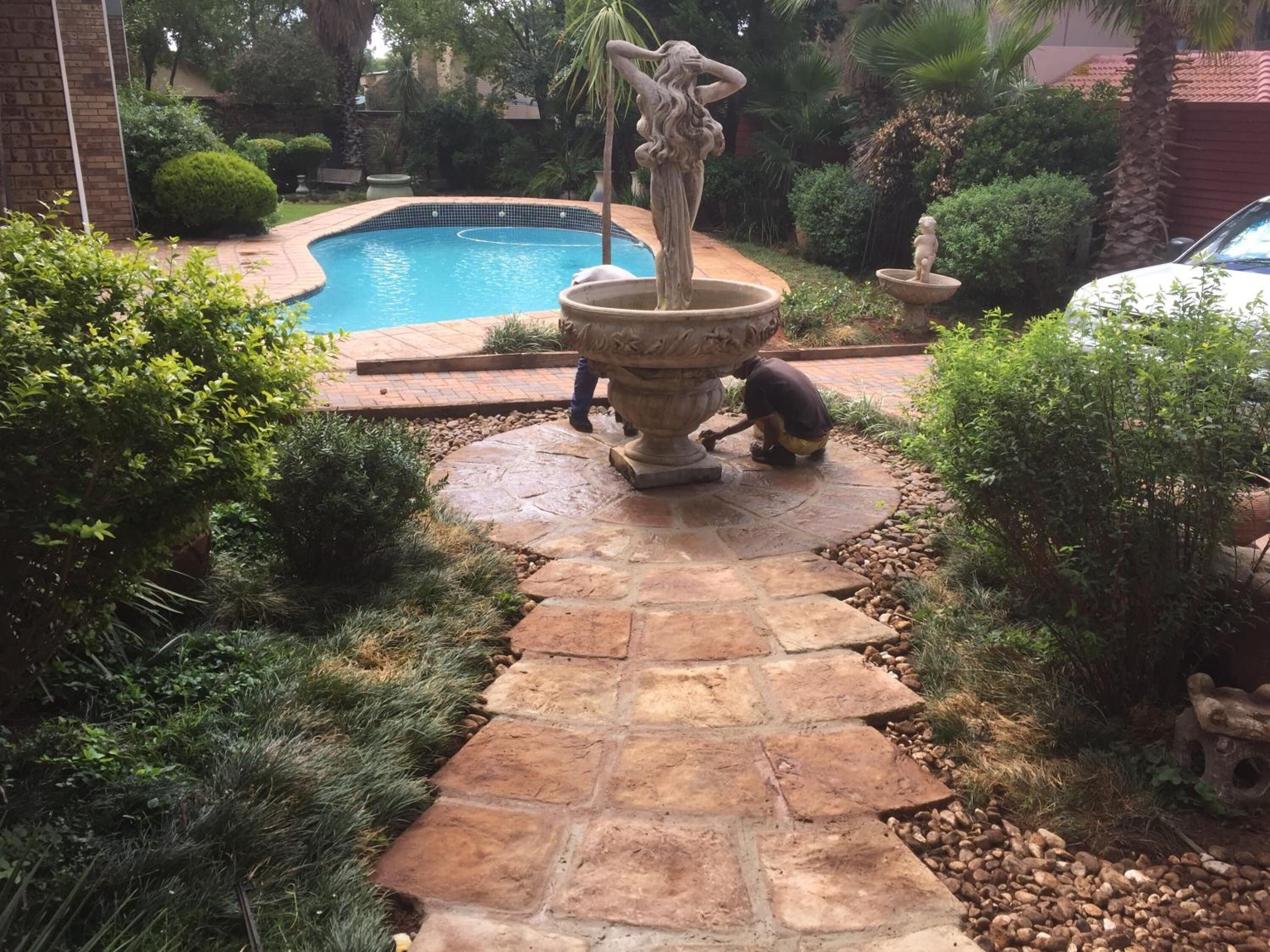 Un sendero de piedra que lleva a una fuente decorativa en un jardín con una piscina al fondo.