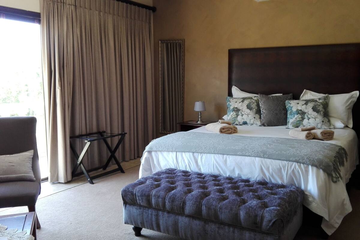 A neatly made bed with a large headboard, two bedside tables with lamps, a tufted ottoman, and a chair.