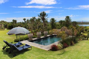 Une piscine dotée de chaises longues et d’un parasol, entourée de plantes tropicales et offrant une vue sur un lac.