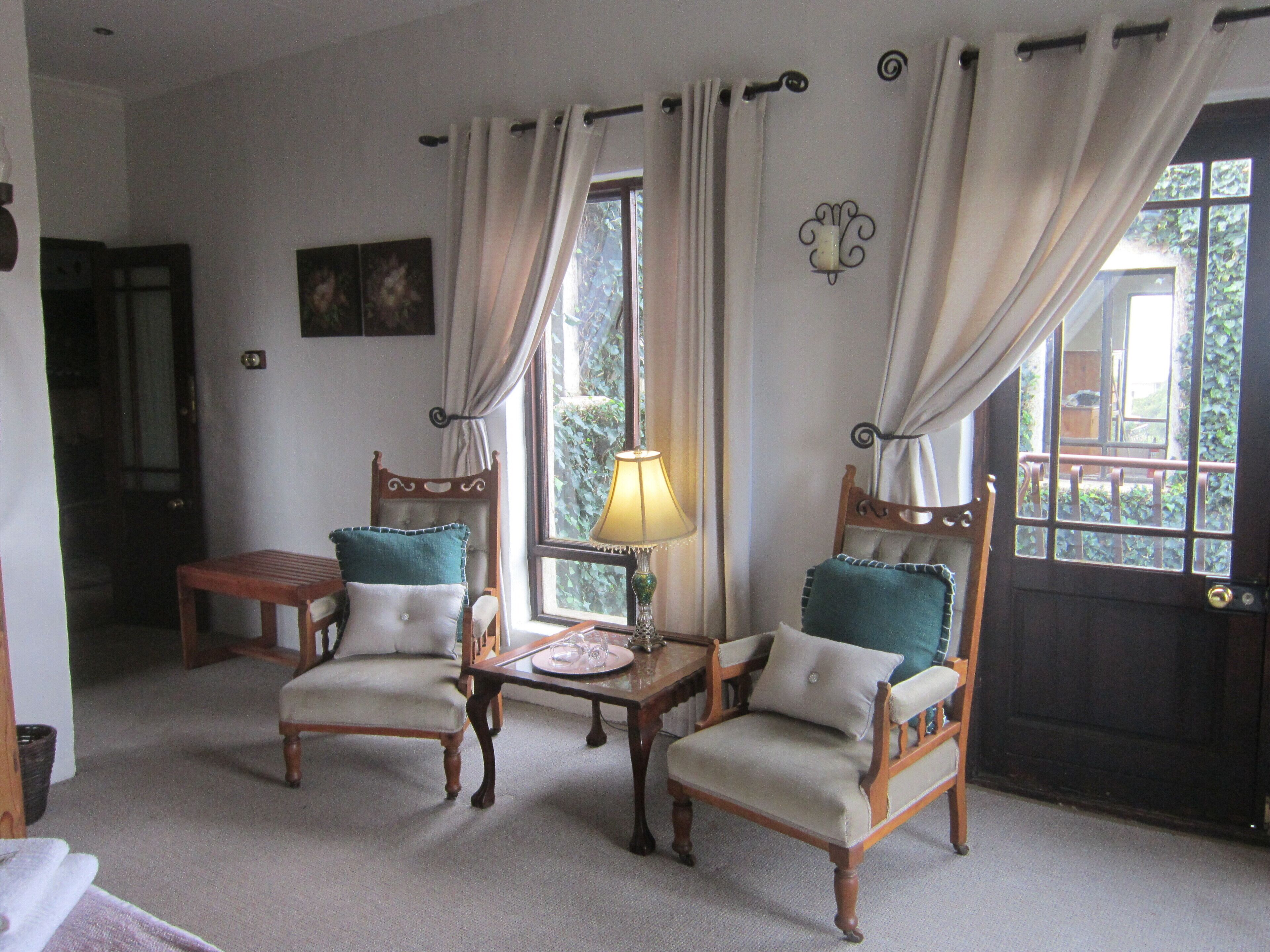 Una habitación con dos sillas de madera con cojines, una mesita con una lámpara y una ventana con cortinas.