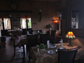 Une salle à manger à la lumière tamisée, avec des tables préparées pour deux personnes, des chaises en osier et une nappe à motifs floraux. Une lampe de table assure l’éclairage, et une plante en pot apporte une touche de verdure.
