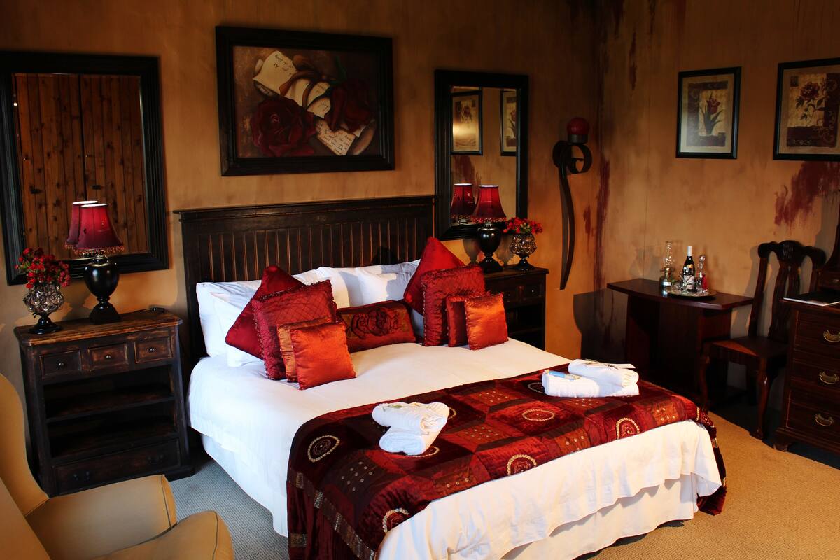 Une chambre d’hôtel avec un grand lit, une literie rouge et blanche, une table de chevet en bois avec une lampe, un bureau avec une chaise et des œuvres d’art encadrées accrochées aux murs.