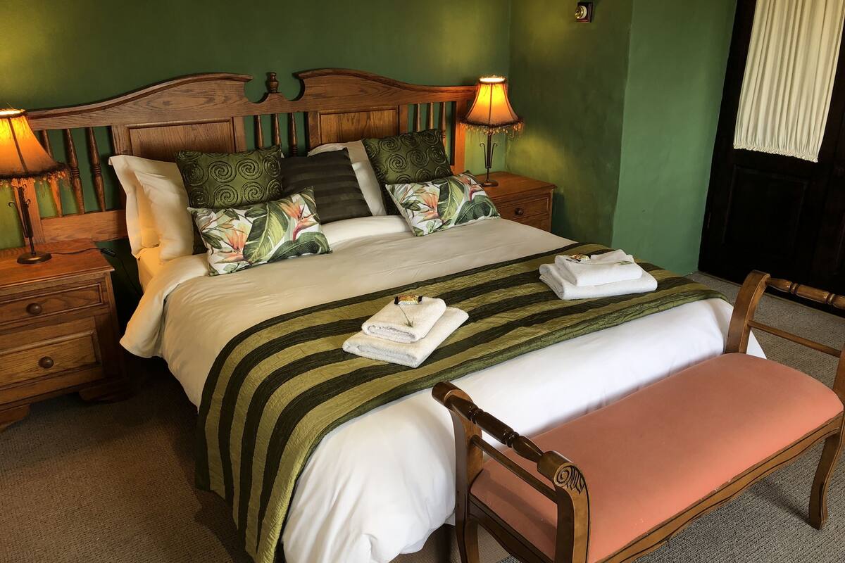 Un lit bien fait, avec une couverture à rayures, des oreillers et une serviette pliée posée sur le côté. On y trouve une table de chevet en bois avec une lampe, ainsi qu’une chaise dotée d’un siège rembourré.