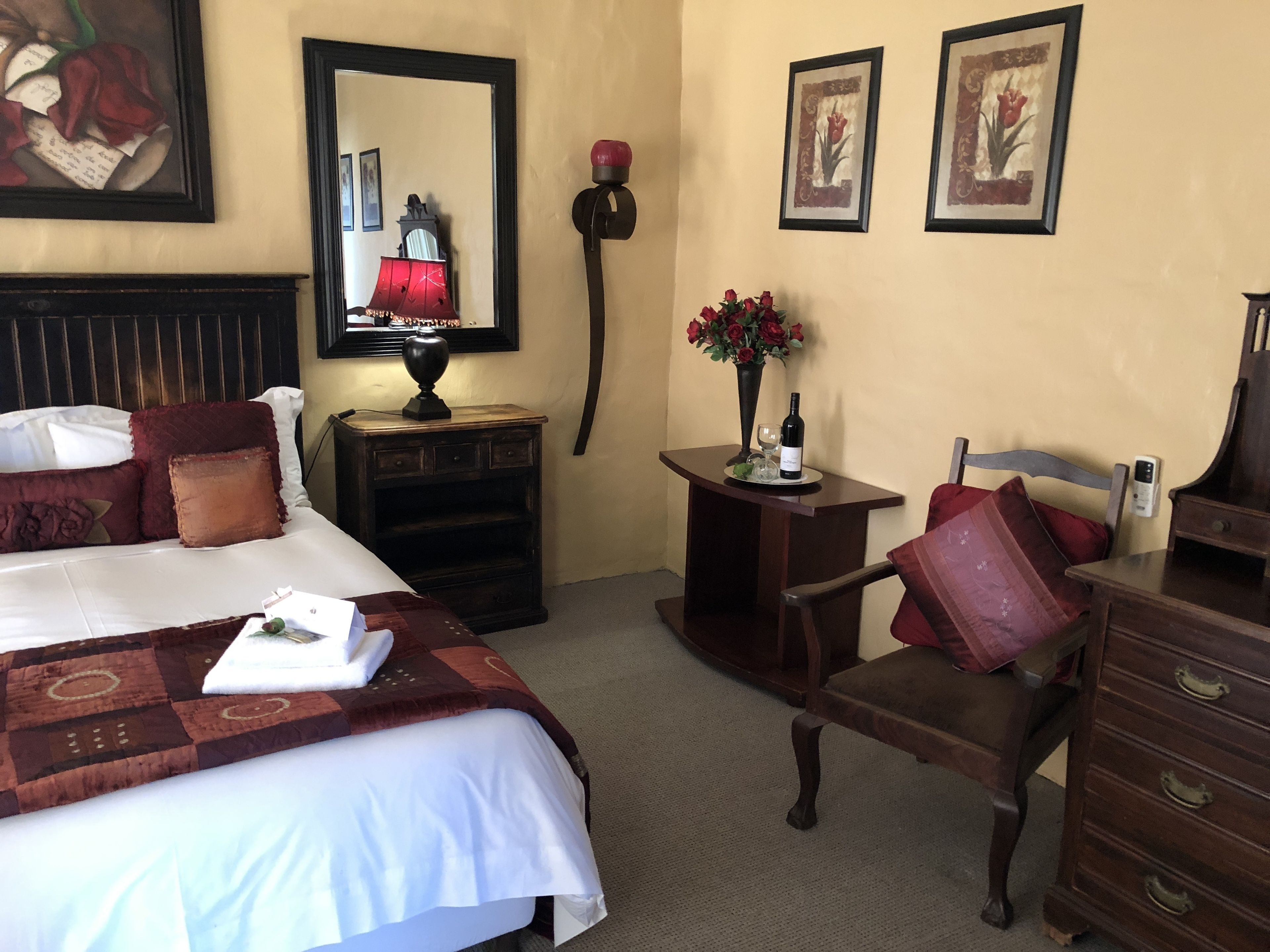 Una habitación de hotel con una cama grande, una mesita de noche con lámpara, una pequeña mesa con un florero y una silla, además de un aparador.