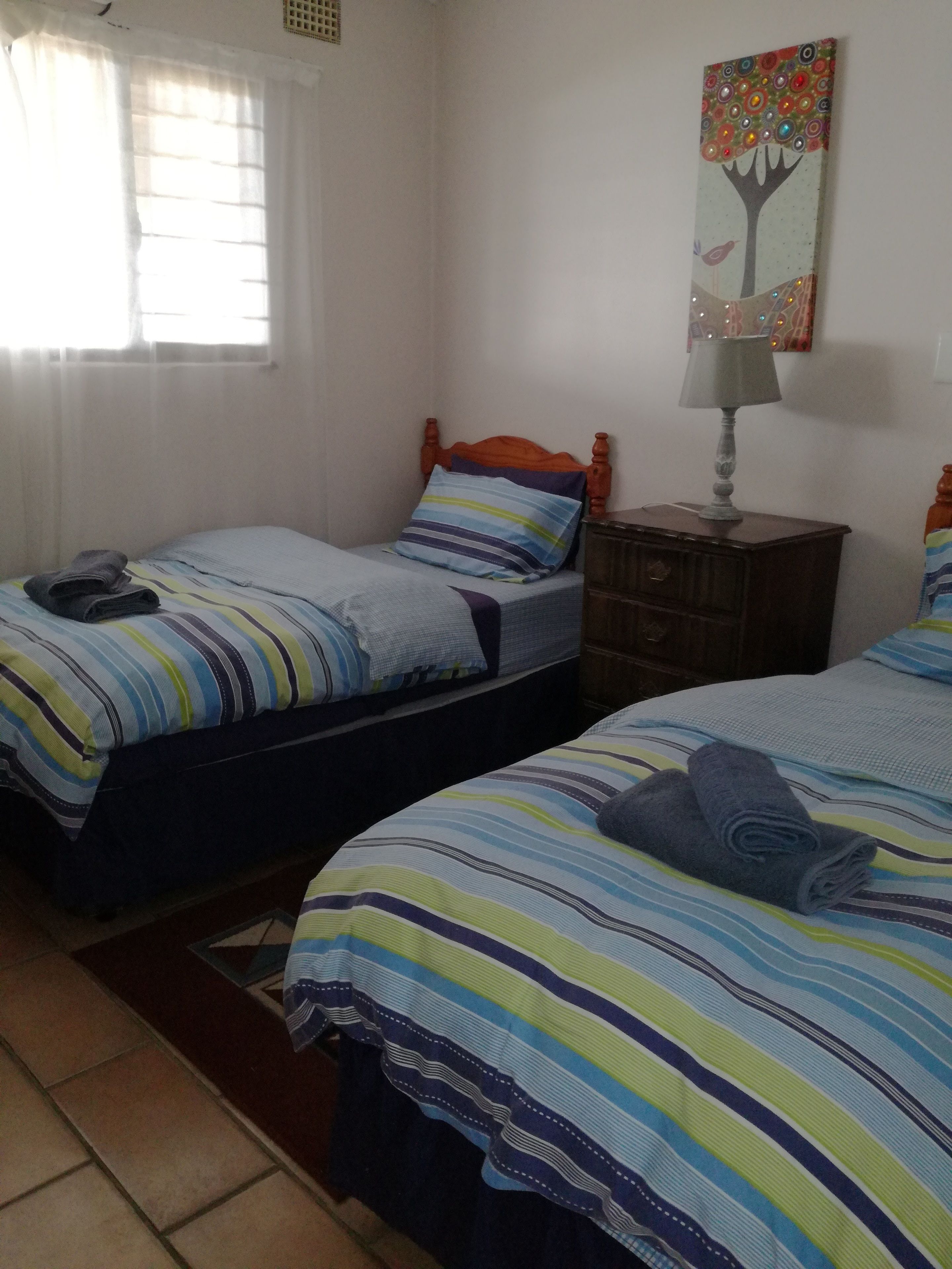 A bedroom with two single beds, a nightstand with a lamp, a window with curtains, and a painting on the wall.