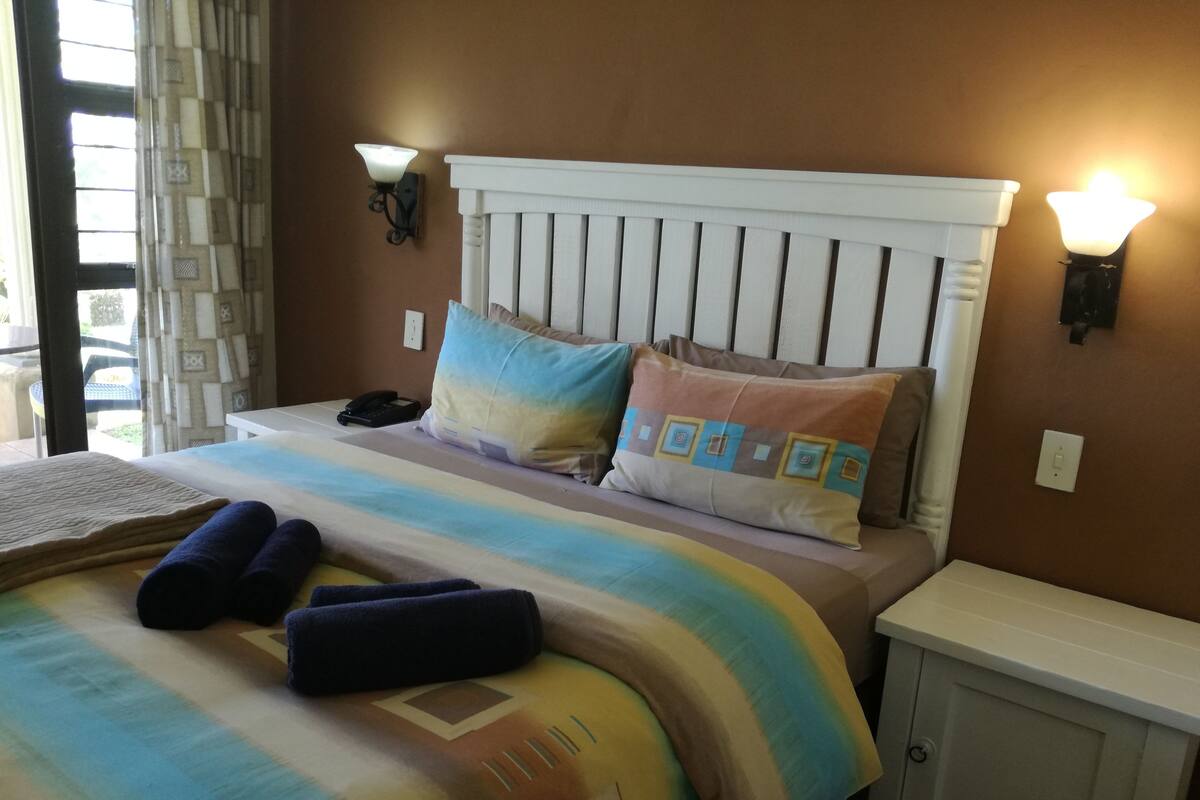A neatly made bed with a striped blanket, two pillows, and rolled towels. There are two wall-mounted lights and a bedside table with a phone.